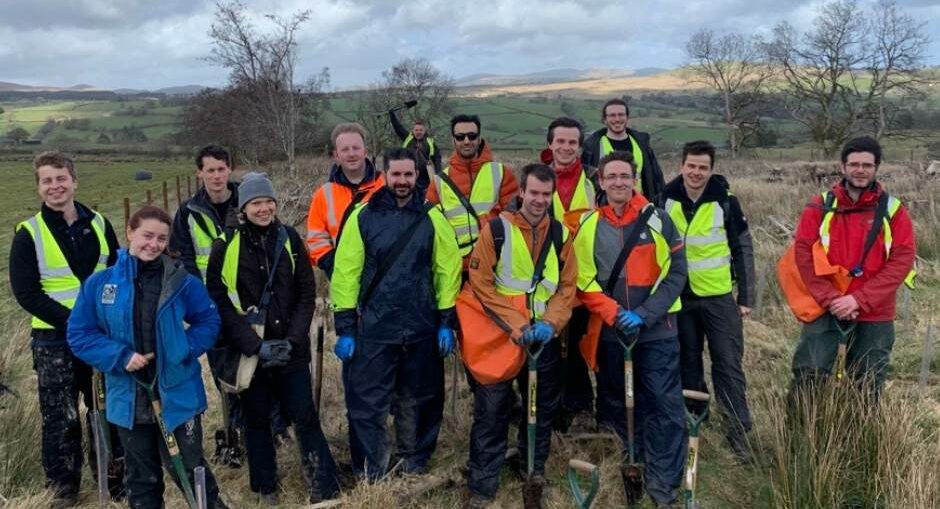 A group of people wearing casual work clothes and gloves are gathered outdoors on an overcast day, smiling and enjoying their time together at a corporate volunteer event focused on conservation.