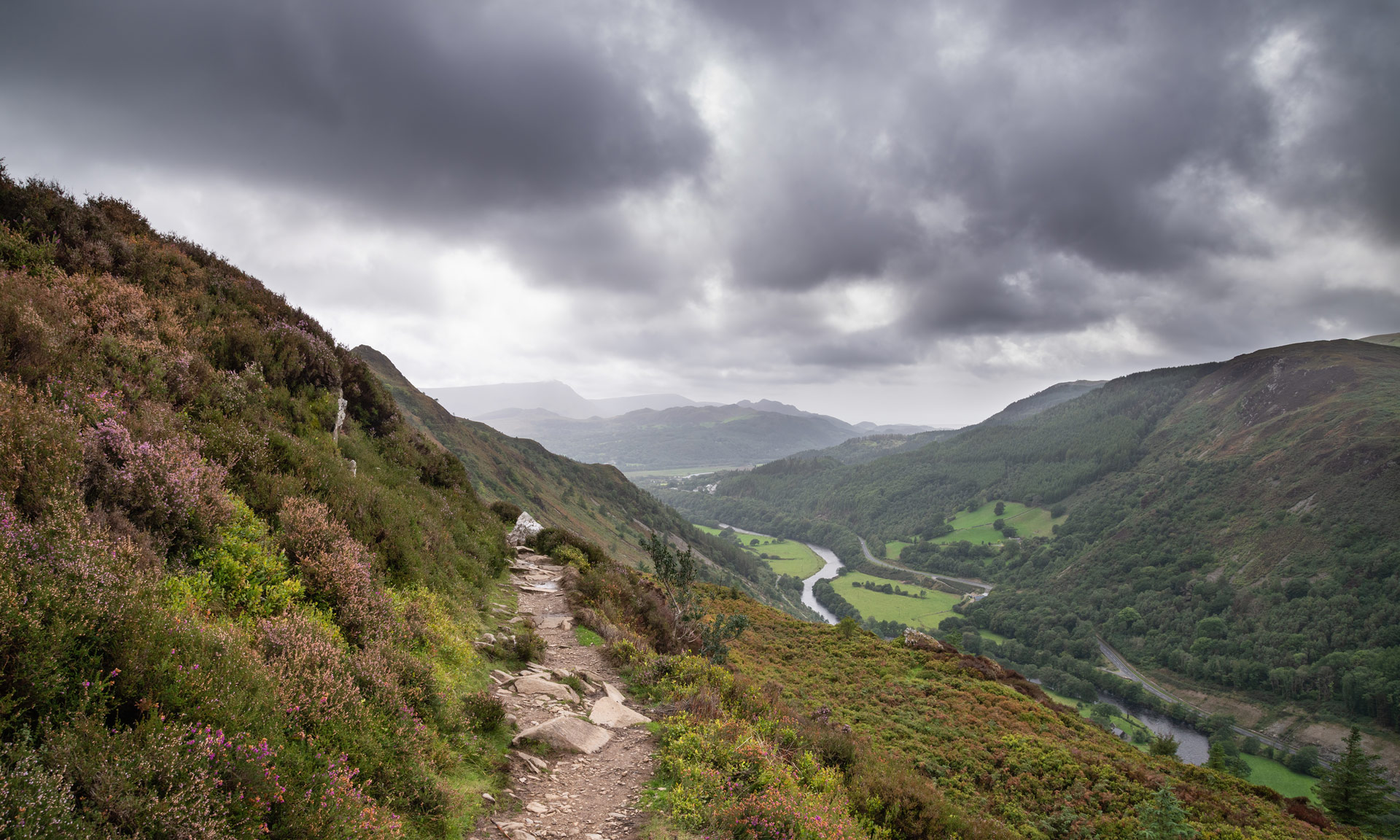 Scenic view from Precipice Walk, showing a narrow path winding along the edge of a steep hillside. The landscape features lush green fields, dense forests, and distant mountains under a clear blue sky. Hikers can be seen enjoying the breathtaking panorama.