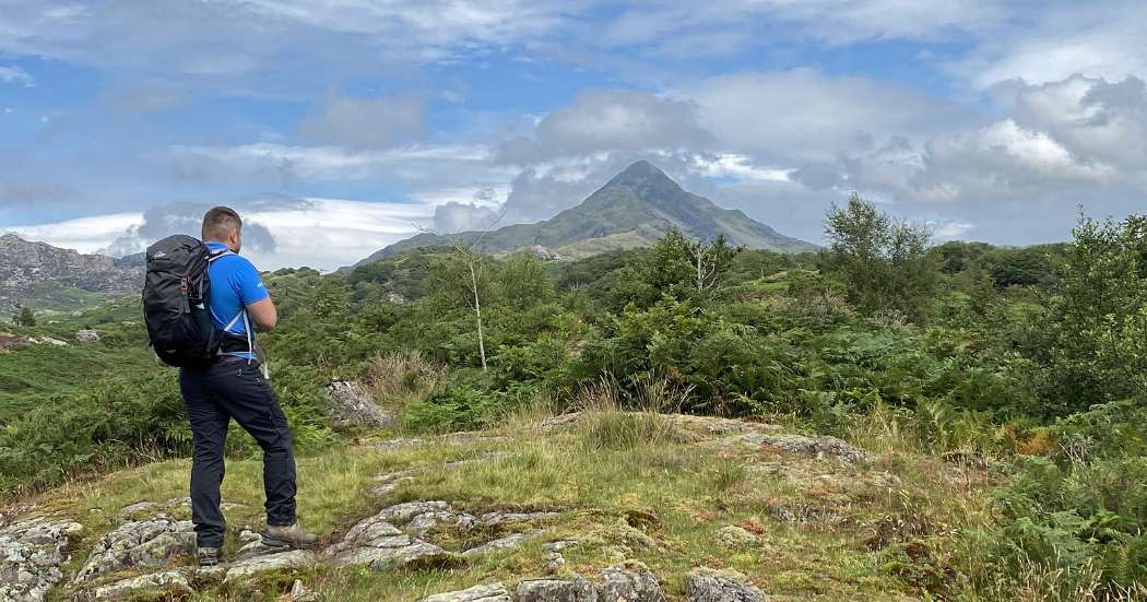 A warden from Eryri National Park stands on rocky terrain, gazing at a cloudy view of Cnicht while on the Croesor circular route. He is wearing a blue polo shirt and a rucksack, blending into the rugged, overcast mountain landscape.